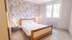 Ein Schlafzimmer mit einem hölzernen Bett, einem Nachttisch mit Lampe, einem Fenster mit Jalousien und einer Wand mit tropischem Blattmuster.