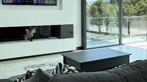 A modern living room with a flat-screen TV mounted on the wall, a black sofa, and a glass balcony with a view of greenery.