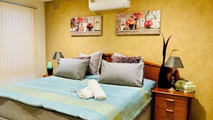Un lit bien fait, recouvert d’une couvre-lit à motifs bleus et verts, avec deux oreillers et une serviette enroulée posée sur le côté. On aperçoit une table de chevet avec une lampe et un petit objet décoratif. Le mur de la pièce est jaune et orné de deux tableaux floraux encadrés.