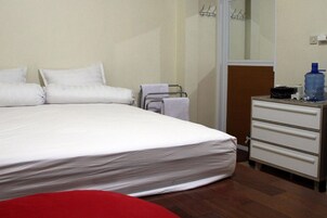 Une chambre d’hôtel avec un grand lit, une commode en bois, une chaise rouge et une bouteille d’eau posée sur la commode.