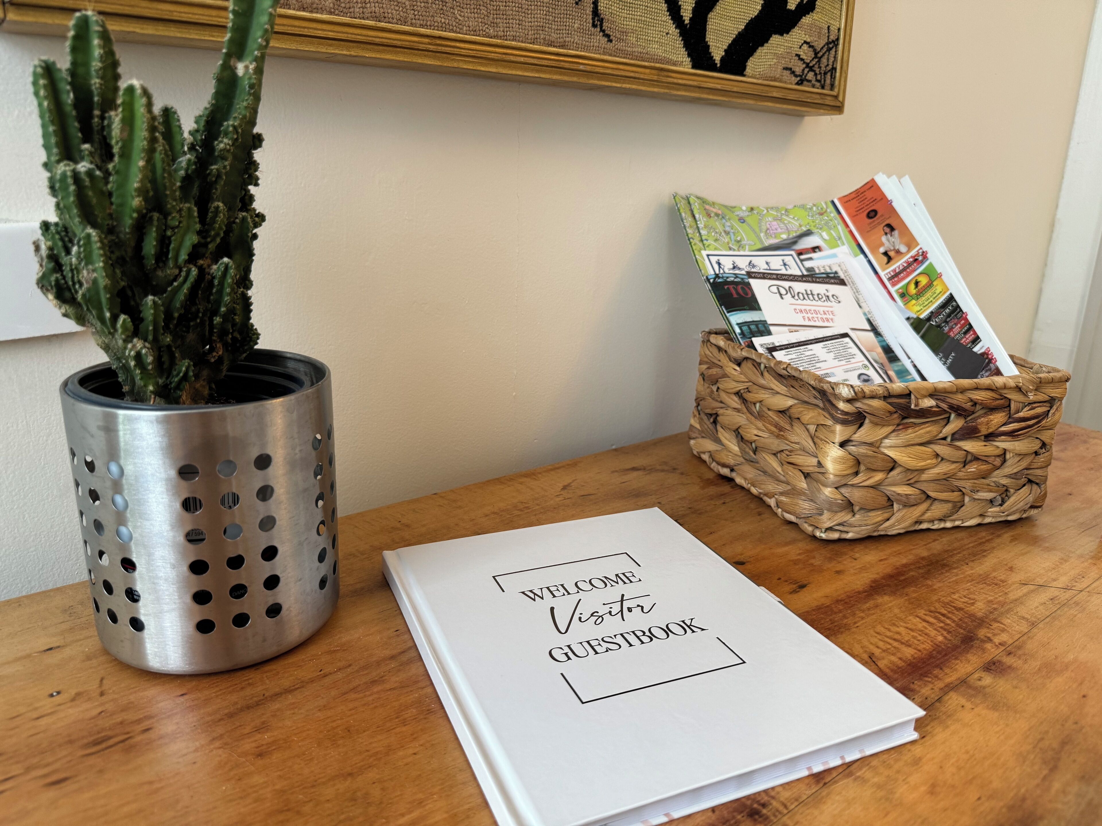 A cactus in a metal pot, a wicker basket with magazines, and a welcome guestbook on a wooden table.