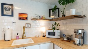 Una cocina moderna con azulejos blancos tipo metro, encimeras de madera, un lavamanos, un microondas, una cafetera y objetos decorativos en estantes.