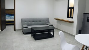 A small, modern room with a grey sofa, a black TV stand, a white chair, and a small table. - MARAZUL-HOME (Playa del Carmen)