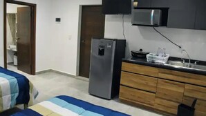 A compact hotel room with a kitchenette, a bed with striped bedding, and a door leading to another room. - MARAZUL-HOME (Playa del Carmen)