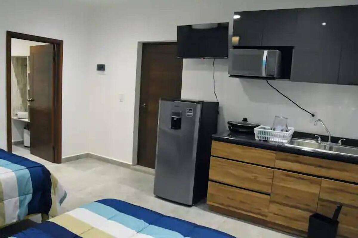 Una habitación de hotel compacta con kitchenette, una cama con ropa de cama a rayas y una puerta que da a otra habitación.