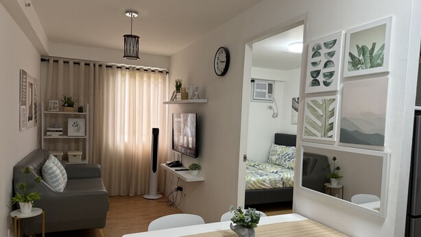 A modern living room with a sofa, a dining table, and a TV. There is a bookshelf with decorative items and a plant. - Cozy & Quiet 1 BR Condo w/ Free Pool Access Near Beach, Airport and Malls (Davao City)
