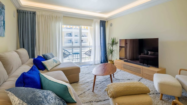 Un salon avec un canapé, un pouf et un téléviseur. Une grande fenêtre, ornée de rideaux, offre une vue sur les bâtiments environnants.