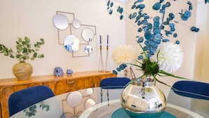 Un coin repas avec une table en verre, des chaises bleues, un vase décoratif avec des fleurs et un meuble en bois doté d’un miroir et de supports à bougies.