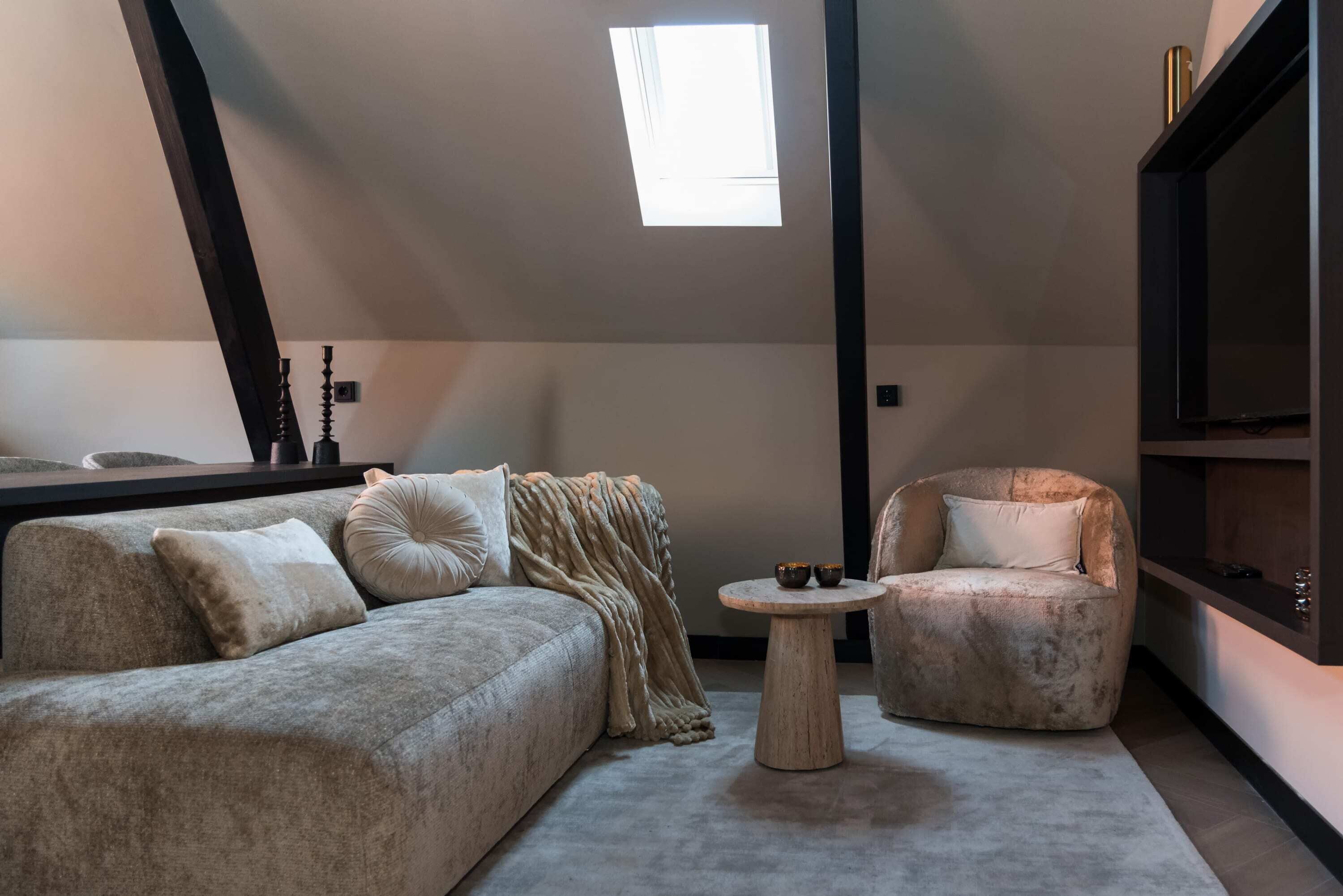 A cozy living room with a sofa, a round coffee table, and a chair. A skylight provides natural light, and a TV is mounted on the wall.