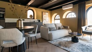 A modern living room with a sofa, chairs, a coffee table, and a decorative vase. There are arched windows and a kitchen in the background.