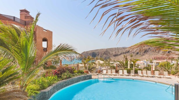 Zona de piscina con sillones de descanso y palmeras, un muro de piedra y vista al mar y las montañas de fondo.