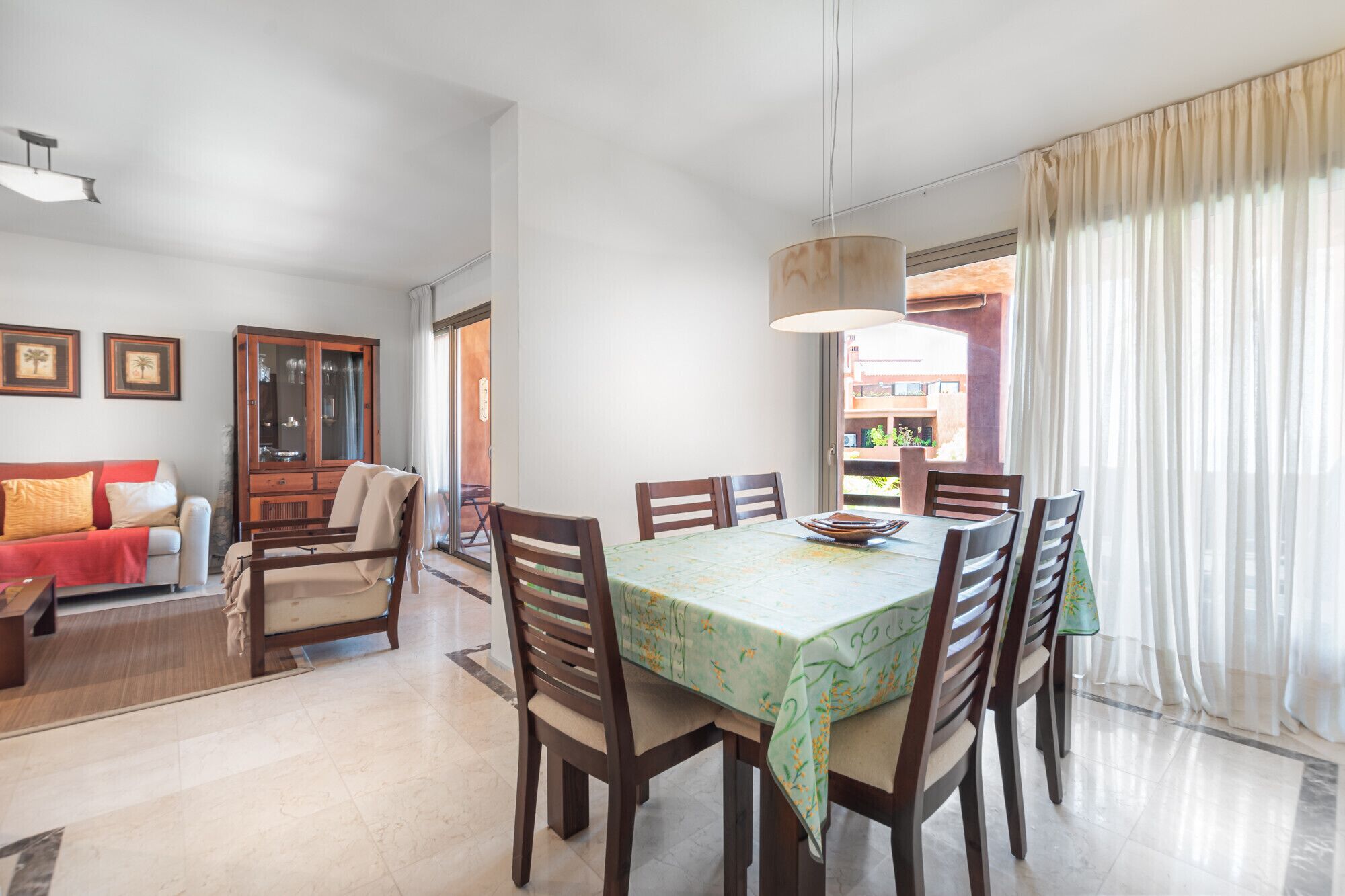 A dining area with a table set for six, surrounded by wooden chairs. There is a sofa and a cabinet in the background.