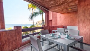 Un comedor con una mesa dispuesta para cuatro personas, con vistas a una playa con palmeras y un litoral rocoso.