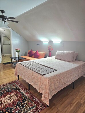 Um quarto com cama, ventilador de teto, mesa de cabeceira, pequena mesa, um vaso com flores, tapete e luminária na parede.