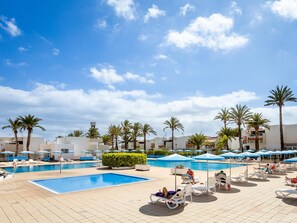 A poolside area with multiple lounge chairs and umbrellas, surrounded by palm trees and buildings.