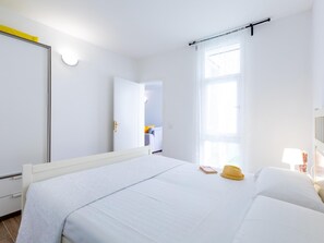 A hotel room with a large bed, a bedside table with a lamp, a window with sheer curtains, and a door leading to another room.