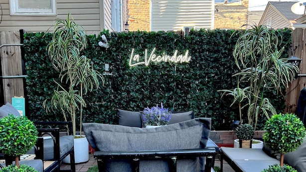 A patio area with a black metal chair, a grey sofa, and a green hedge with a neon sign reading "La Vecindad".