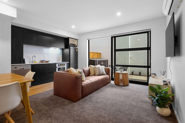 A modern apartment with a kitchen, a living area with a sofa, a dining table, and a large window.