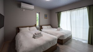 Ein Hotelzimmer mit zwei Betten, einem Flachbildfernseher, einem Fenster mit durchsichtigen Vorhängen und einer wandmontierten Klimaanlage.