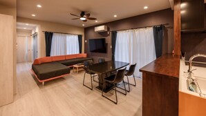 Ein modernes Wohnzimmer mit einer Couch, einem Esstisch und Stühlen. Es gibt einen Deckenventilator, einen an der Wand montierten Flachbildfernseher und eine Küchenzeile mit Spüle und Arbeitsplatte.