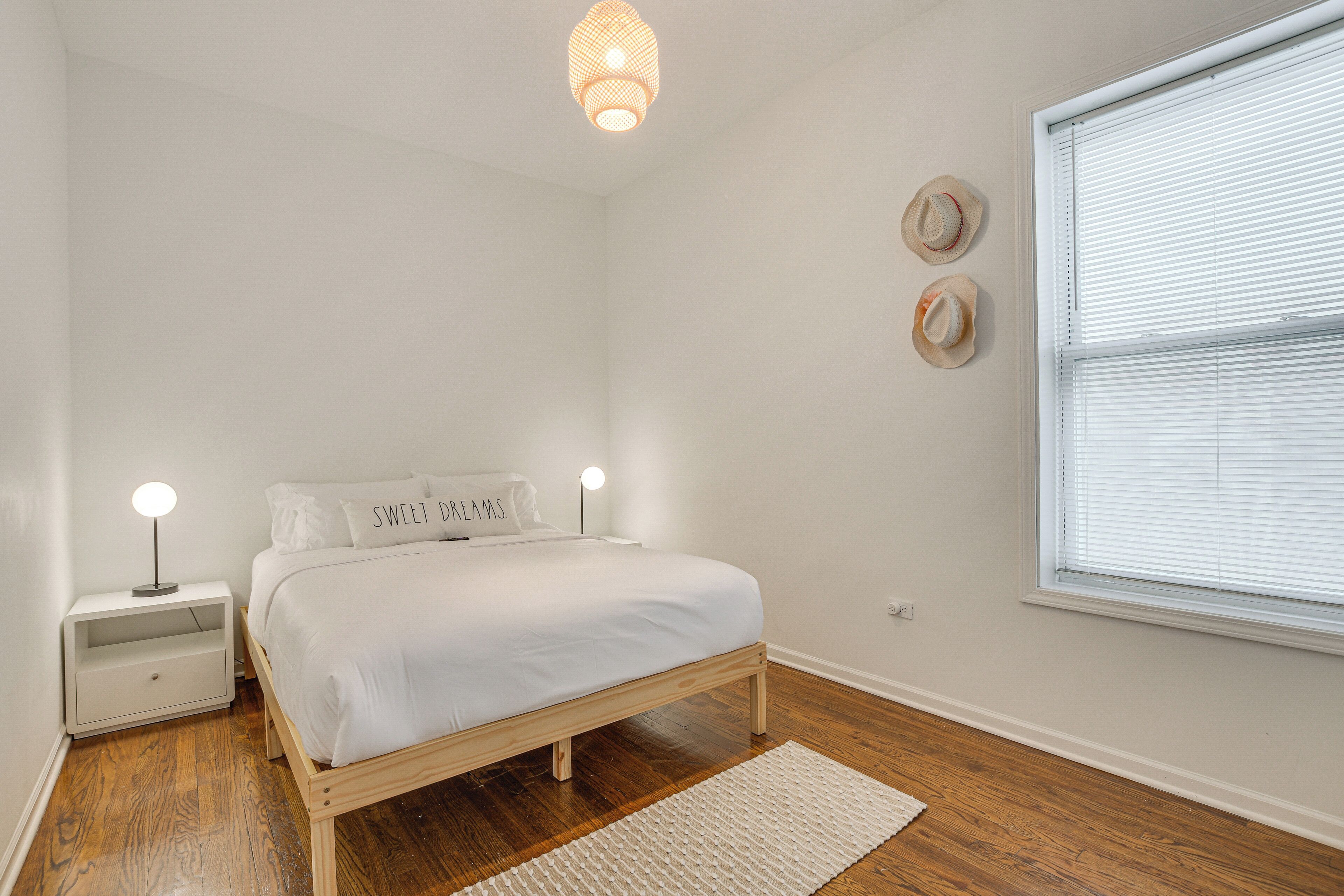 A bedroom with a wooden bed, white bedding, a nightstand with a lamp, and a window with blinds.