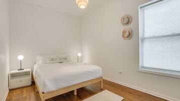 A bedroom with a wooden bed, white bedding, a nightstand with a lamp, and a window with blinds.
