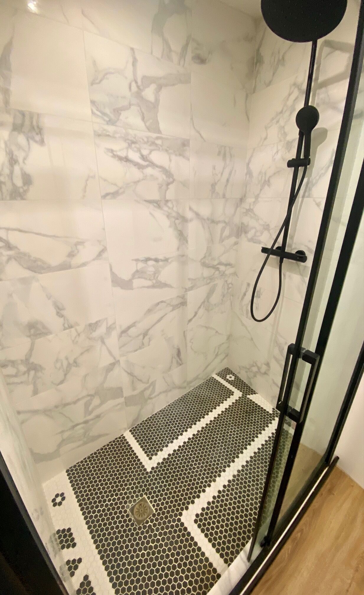 A shower with a glass door, a marble wall, and a tiled floor with a geometric pattern.