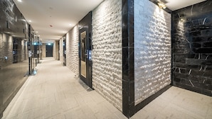 A modern hotel corridor with textured walls, a carpeted floor, and a glass elevator.