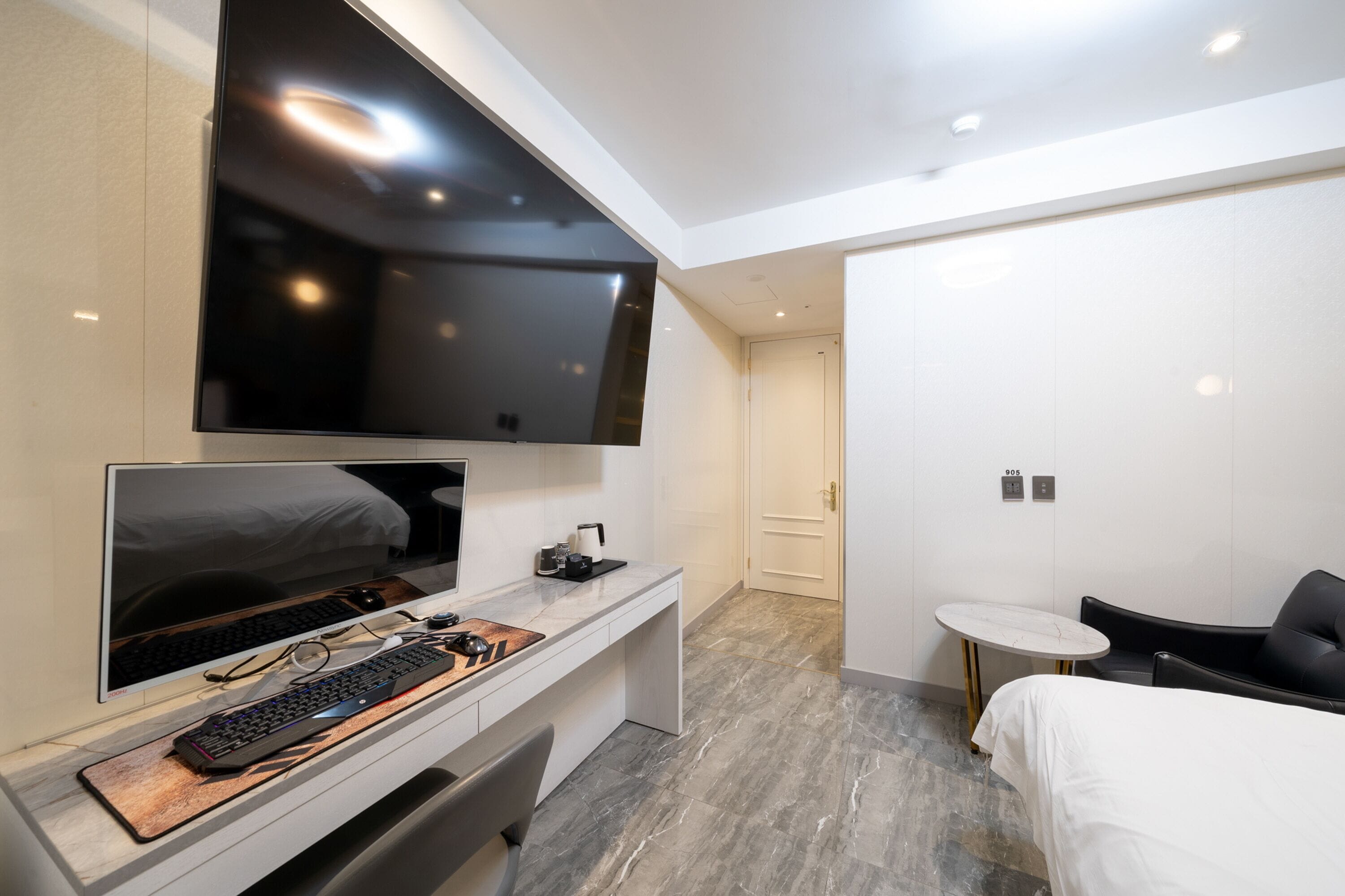 A modern hotel room with a large flat-screen TV mounted above a desk, a bed with a white comforter, a small round table, and a black leather chair.