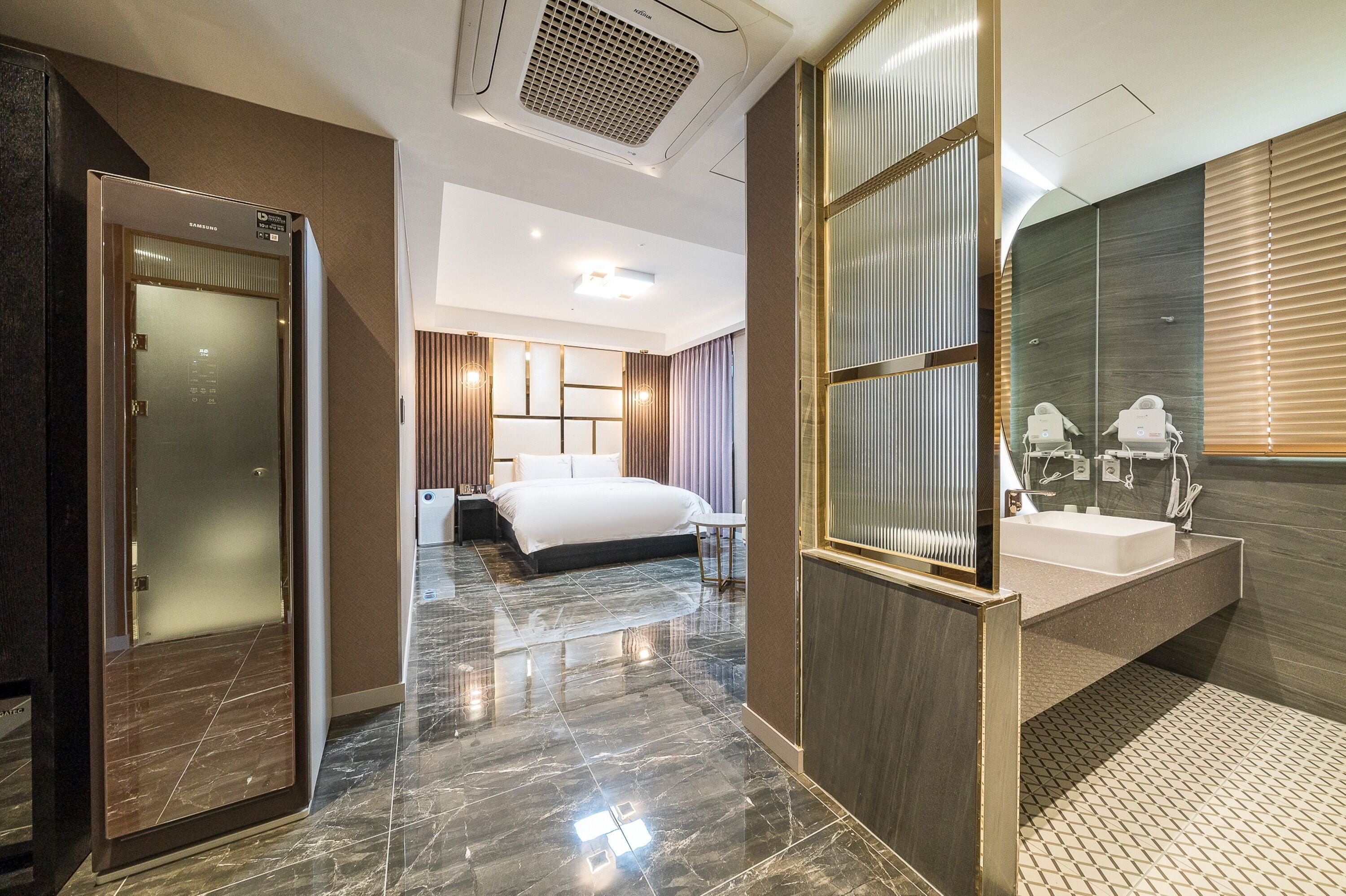 A modern hotel room with a marble floor, a large bed, a separate bathroom with a large mirror and sink, and a separate bedroom with a desk and chair.