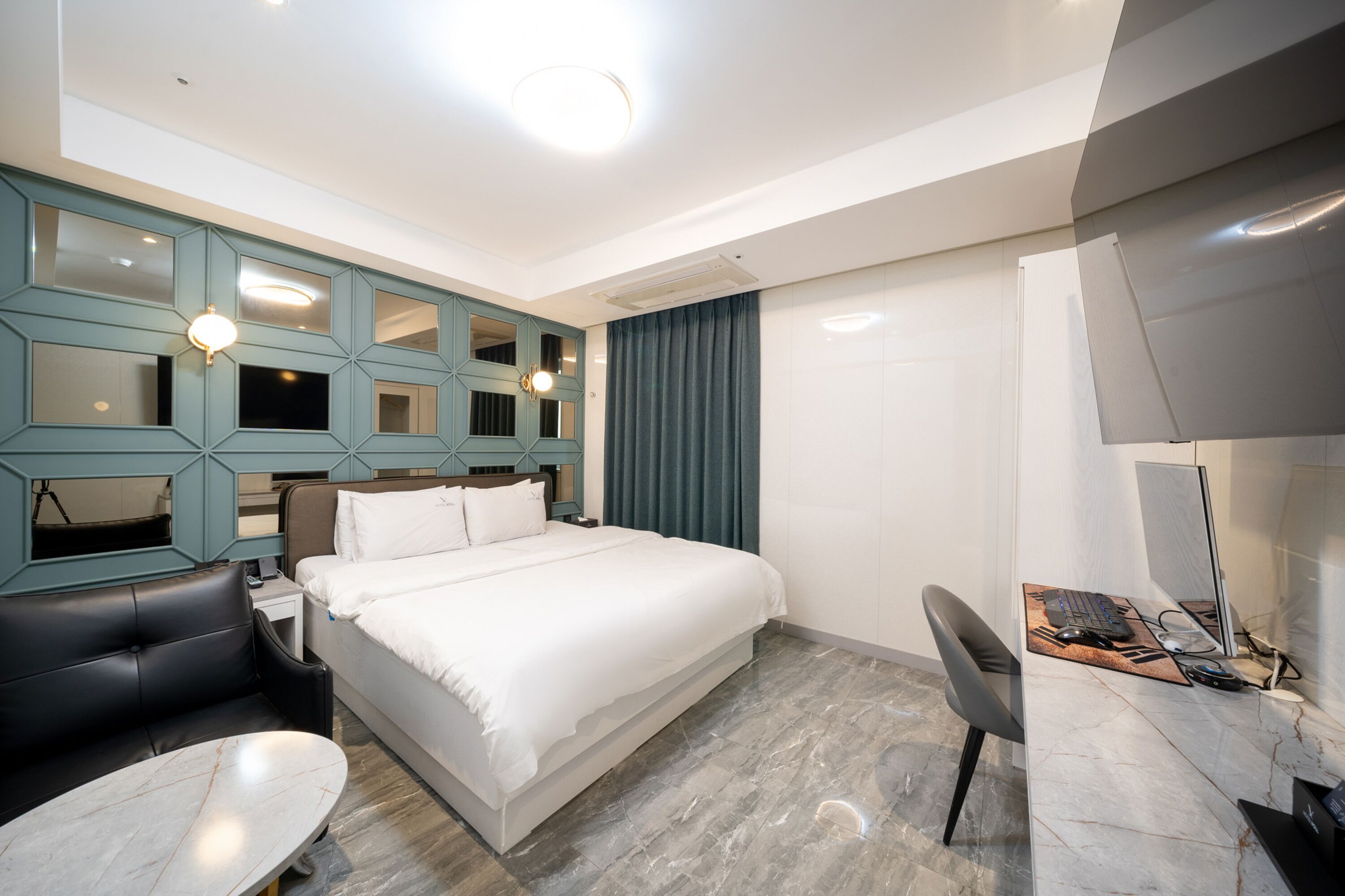A modern hotel room with a large bed, a desk with a computer, a chair, a small table, and a blue accent wall with mirrors.