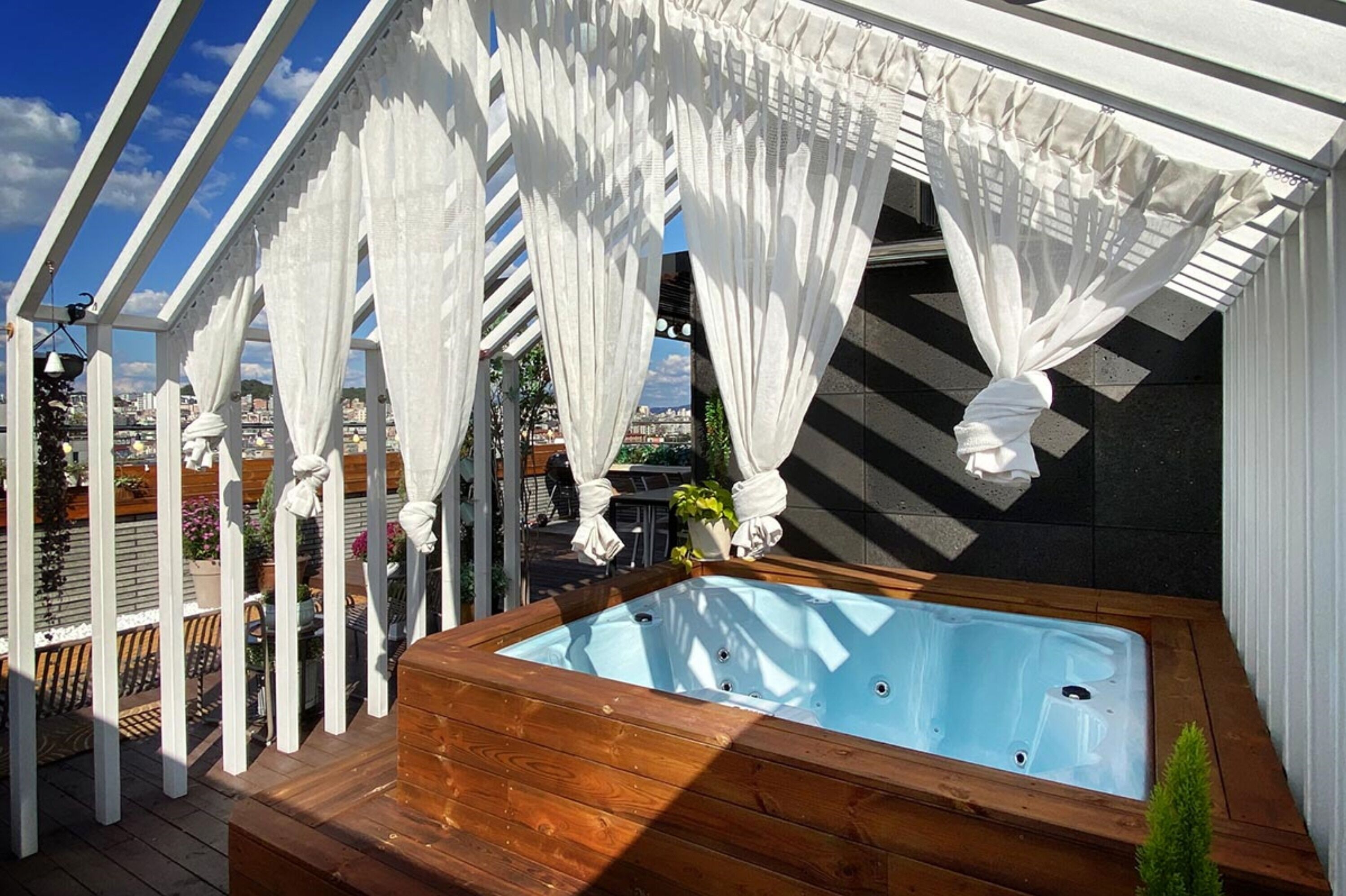 A wooden deck with a hot tub, white curtains, and a view of the city.