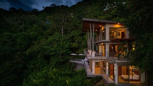 Una casa moderna de varios pisos con puertas y balcones de vidrio, rodeada por un bosque denso.
