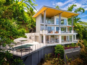 A modern two-story house with a balcony, a swimming pool, and lush greenery surrounding it.
