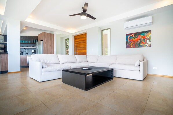 A modern living room with a sectional sofa, a black coffee table, a ceiling fan, and an air conditioning unit.