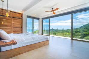 Une chambre moderne avec un grand lit, une tête de lit en bois et une vue imprenable sur un paysage luxuriant grâce à de grandes fenêtres du sol au plafond.