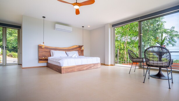 Un dormitorio moderno con una cama de madera, un ventilador de techo y vistas a zonas verdes a través de amplios ventanales.