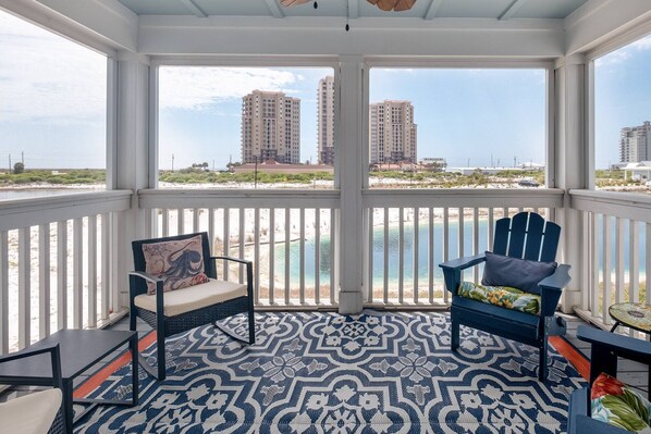 A covered balcony with two chairs, a patterned floor, and a view of the beach and buildings.