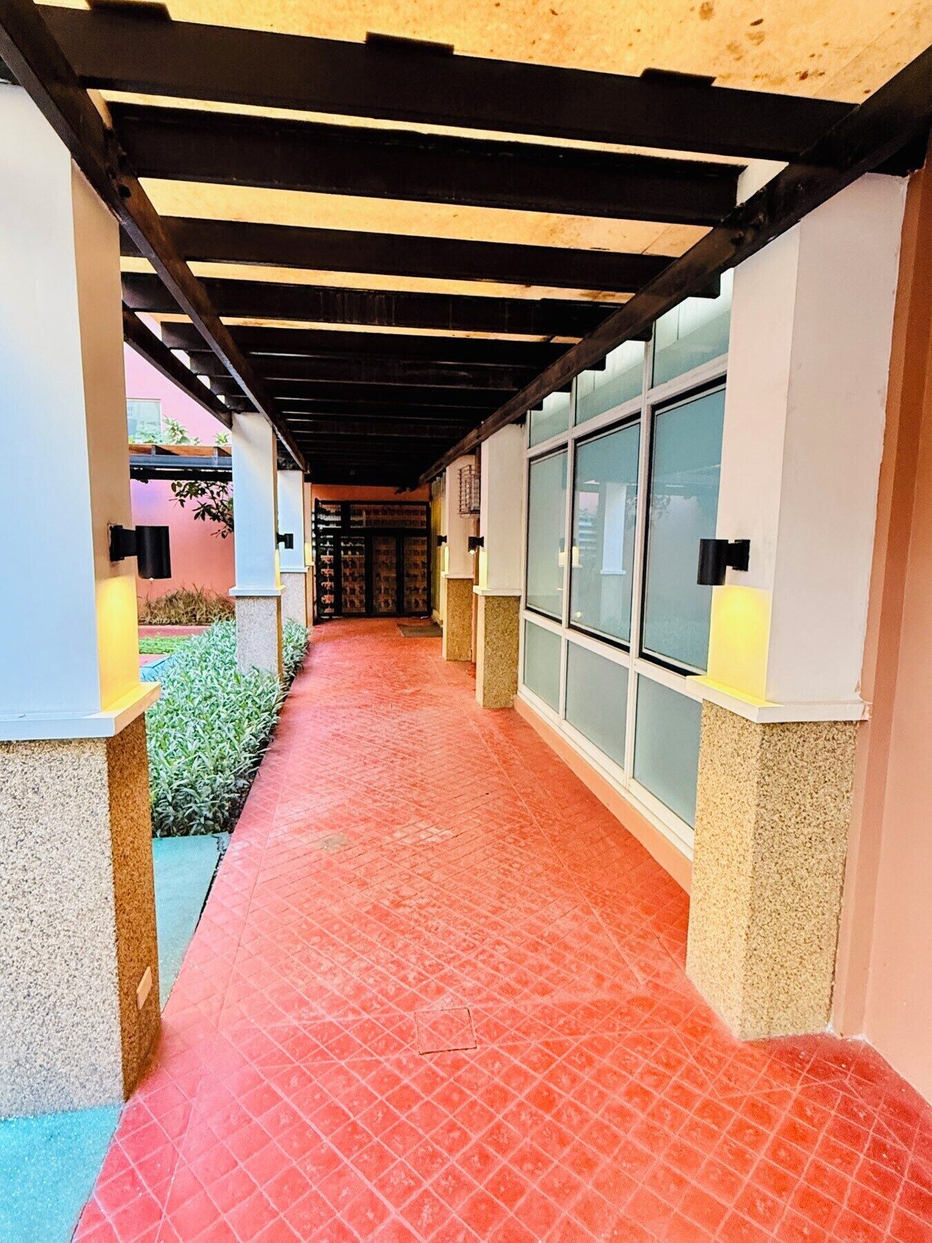 Une allée couverte dotée d’un sol carrelé de tuiles rouges, de murs blancs et de portes vitrées donnant accès à un bâtiment.