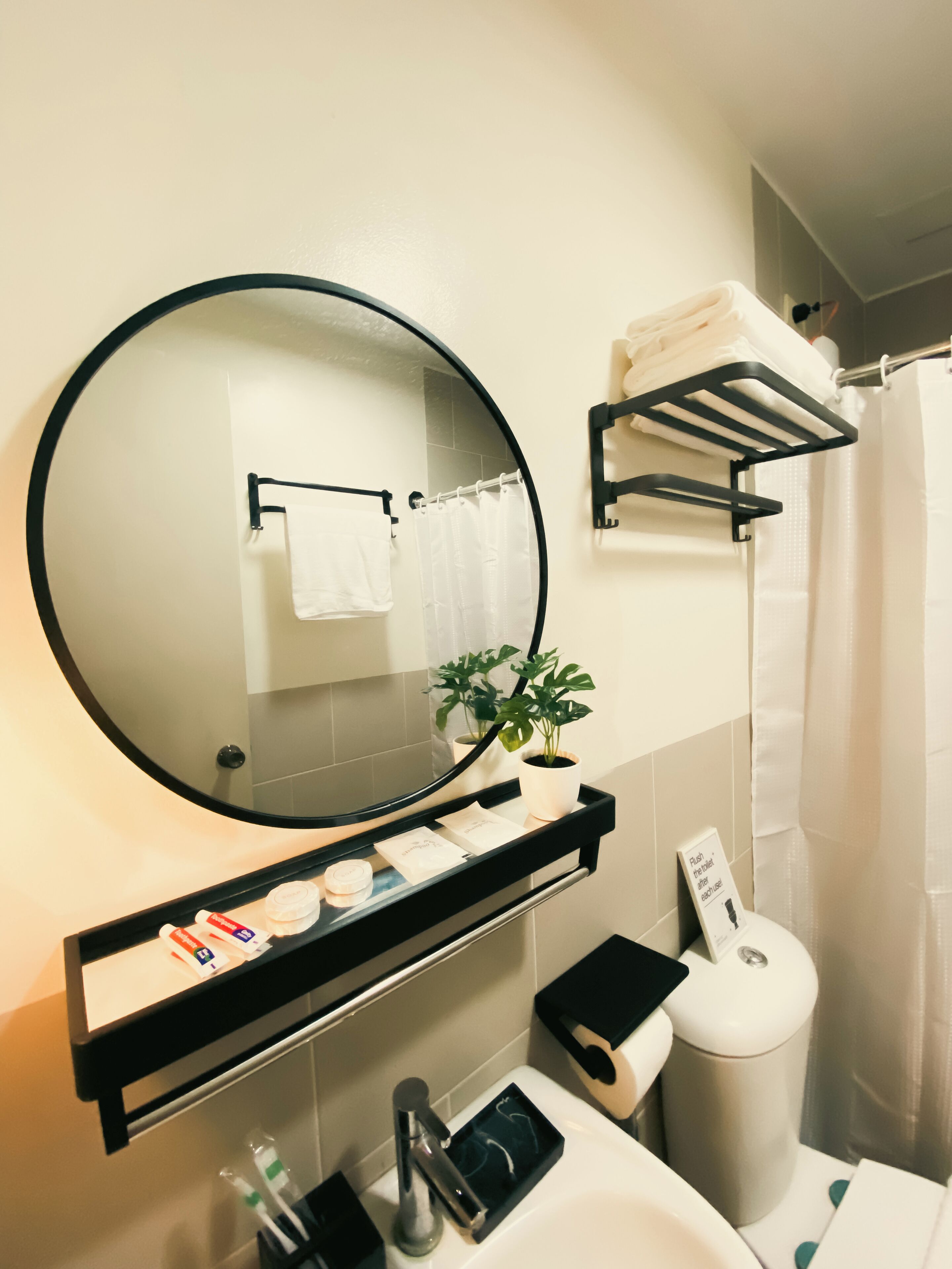 A bathroom with a round mirror, a sink, a towel rack, a shelf with toiletries, a potted plant, and a shower curtain.