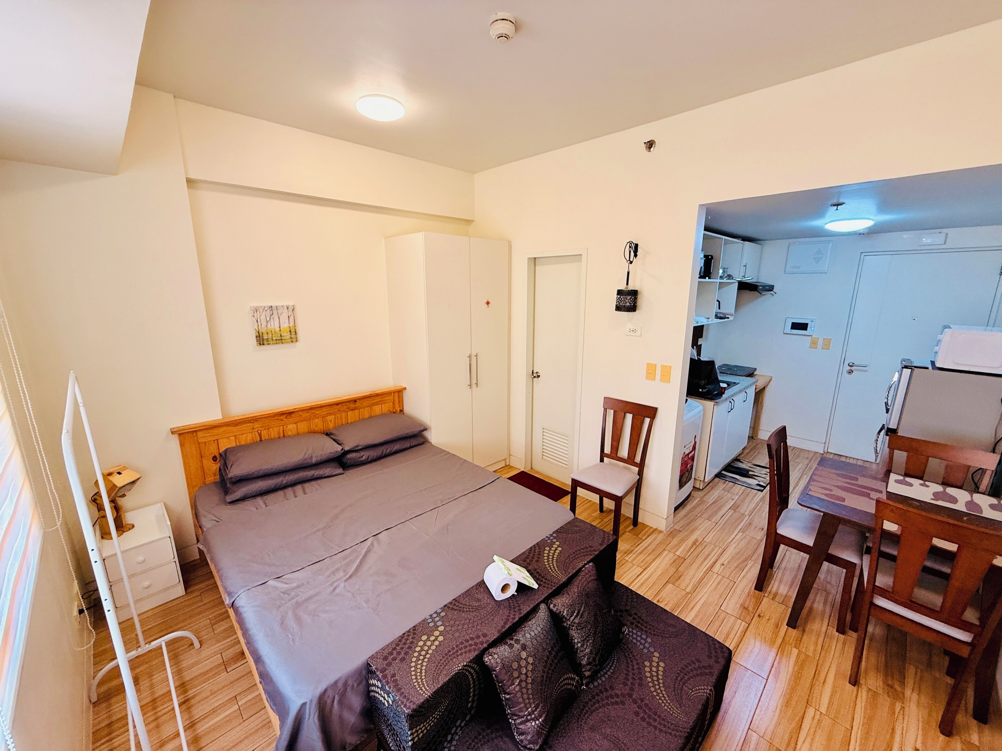 A compact room with a bed, a small dining table, a chair, and a kitchenette.