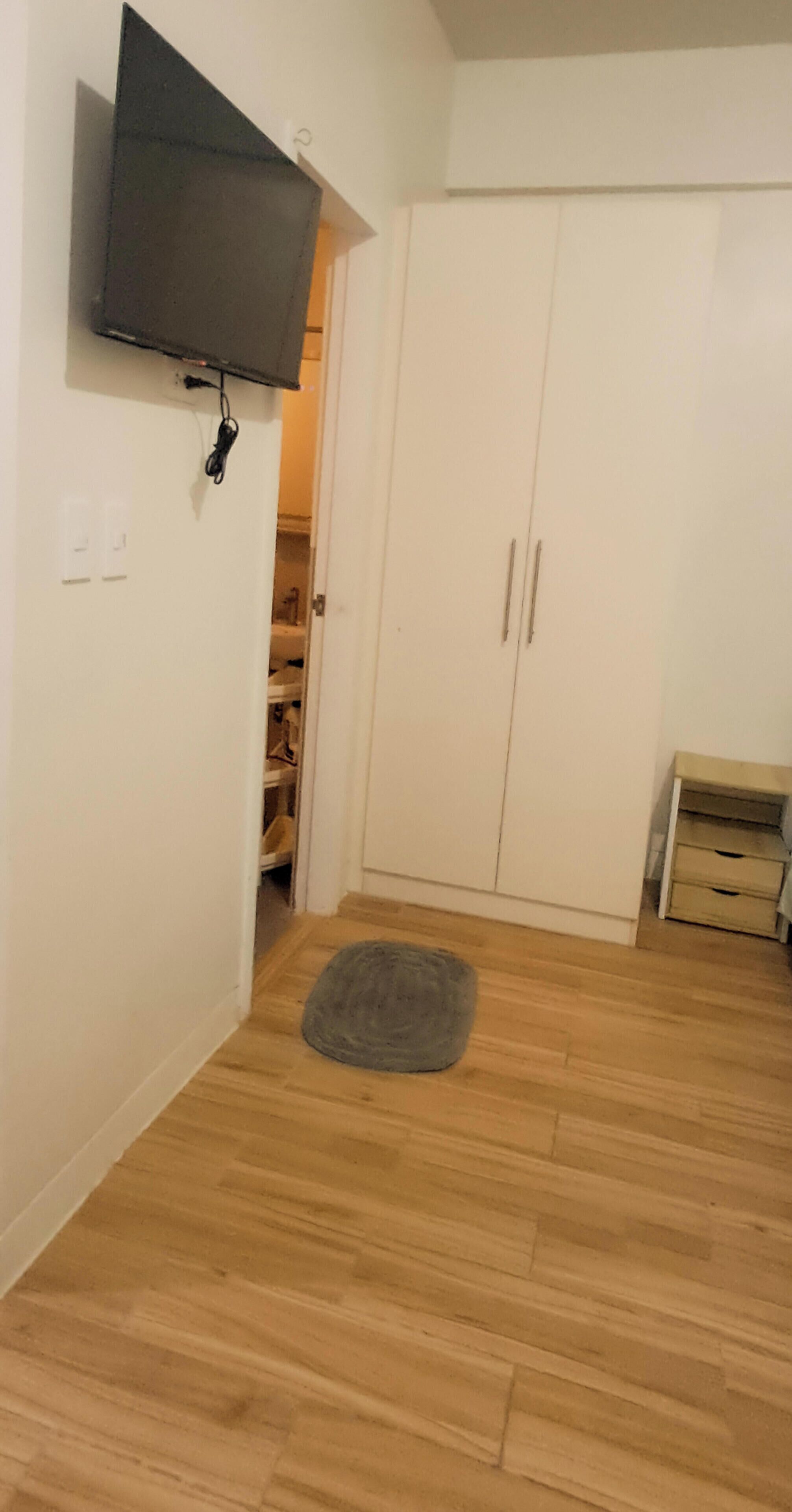 A room with a wooden floor, a grey rug, a wardrobe, and a TV mounted on the wall.