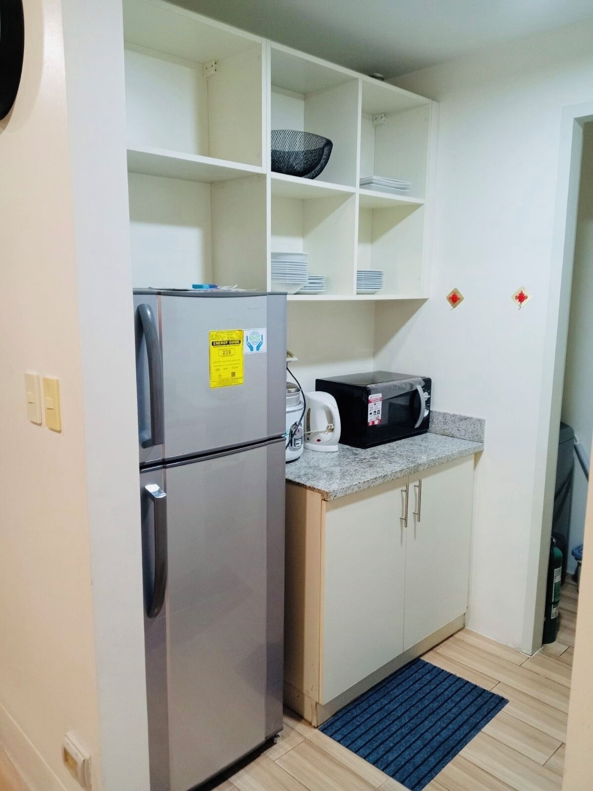 A compact kitchen with a stainless steel refrigerator, a microwave, and a coffee maker on the countertop. There are white cabinets and a blue mat at the entrance.