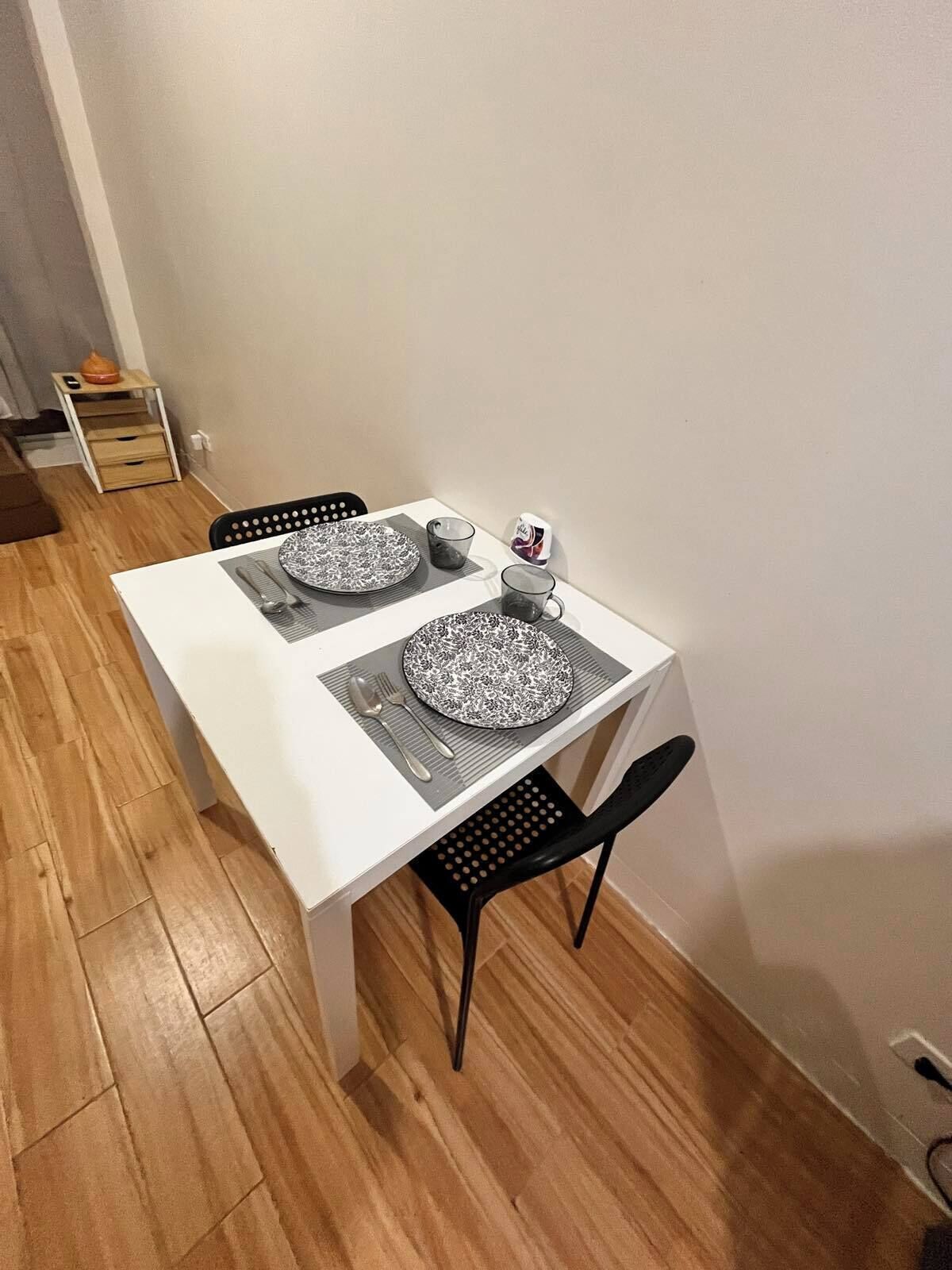 Un petit coin repas avec une table blanche et deux chaises noires. On y trouve deux assiettes et deux tasses.