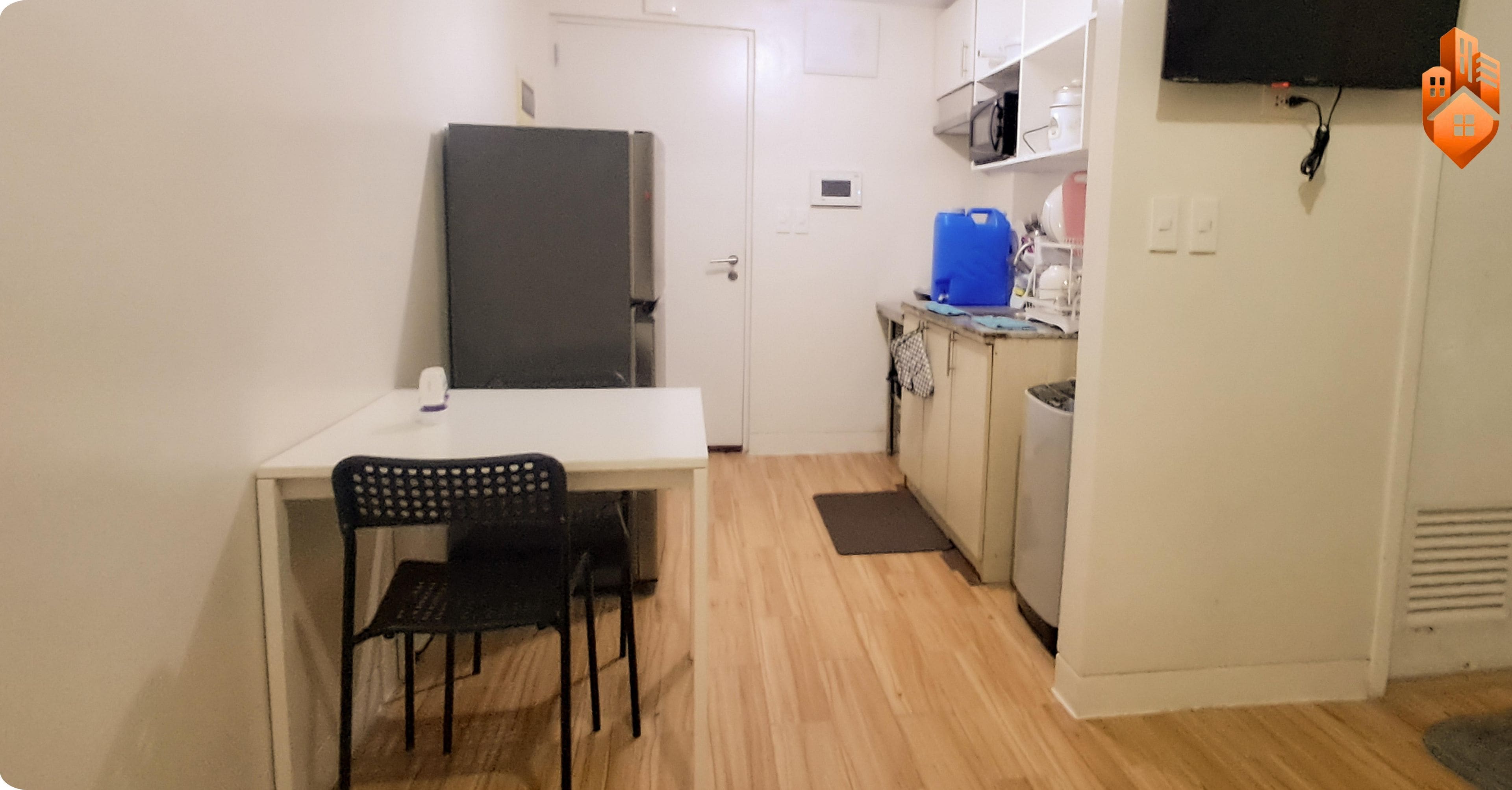 A compact kitchen with a refrigerator, microwave, and sink. There is a small dining area with a chair and a table. The room has white walls and a wooden floor.