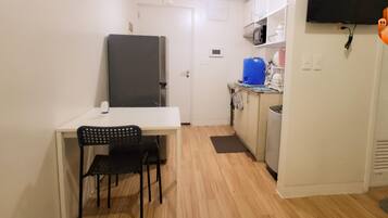 A compact kitchen with a refrigerator, microwave, and sink. There is a small dining area with a chair and a table. The room has white walls and a wooden floor.
