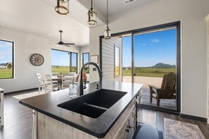 Una cocina moderna con una isla grande, un comedor con sillas, un reloj en la pared y vistas al exterior a través de amplios ventanales corredizos.