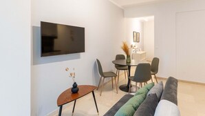 A modern living room with a flat-screen TV mounted on the wall, a dining area with a round table and chairs, and a sofa with multiple cushions. - Phaedrus Living: Alimos Edem Beach Flat (Alimos)