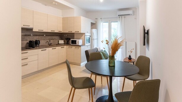 A modern kitchen with light-colored cabinets, a round dining table with chairs, and a vase with dried plants on the table. - Phaedrus Living: Alimos Edem Beach Flat (Alimos)