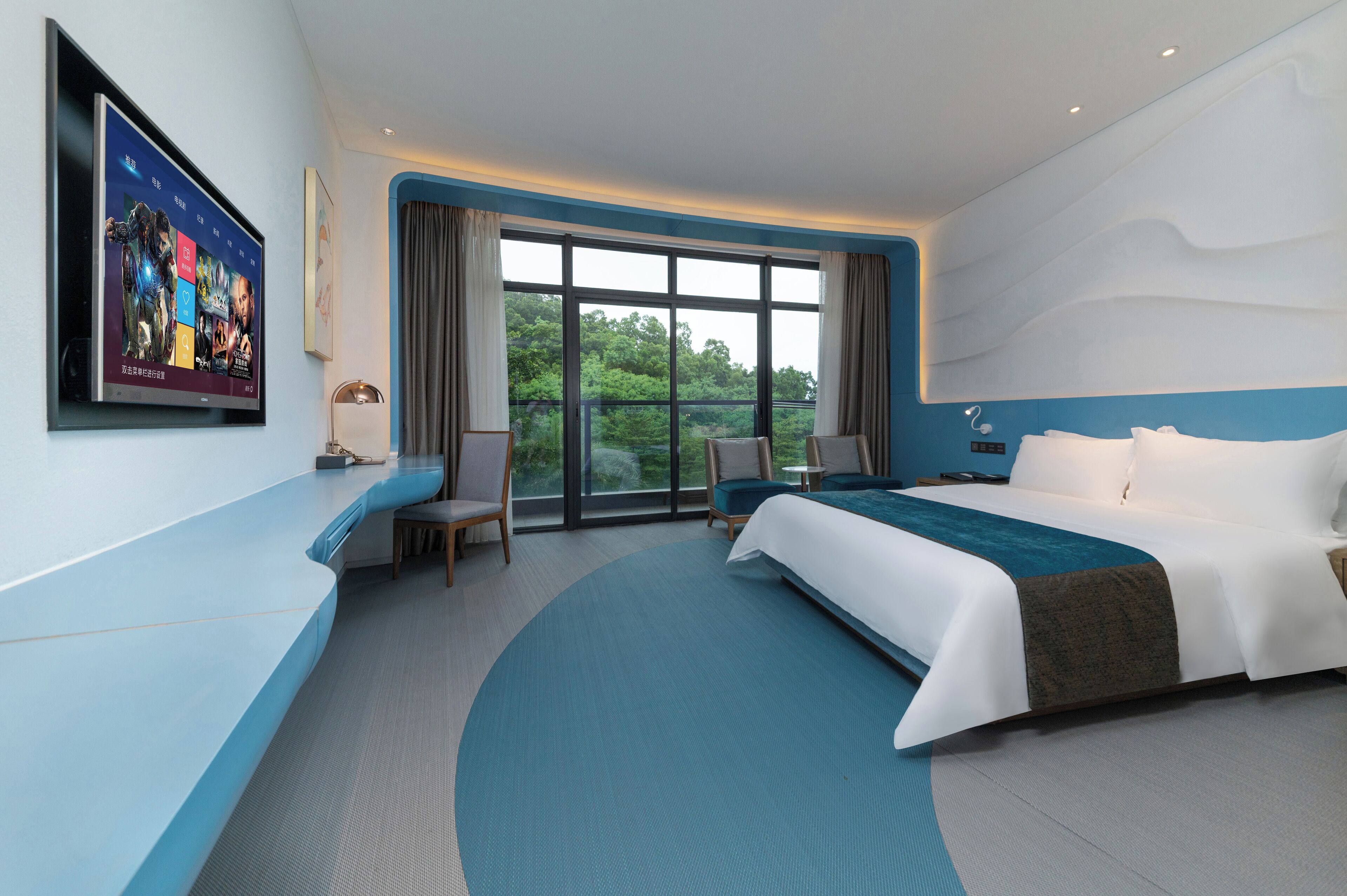 A modern hotel room with a large bed, a seating area with a chair and a small table, a large window with curtains, and a flat-screen TV mounted on the wall.